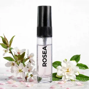CocoBerry Rosea 5ml tester perfume bottle on a light surface with delicate blossom branches, a white gardenia, and scattered pink petals.